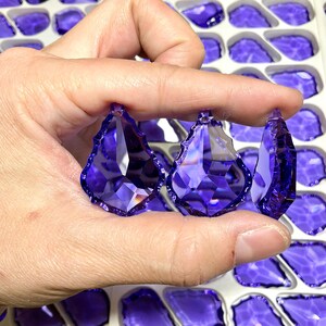 May include: Three faceted, teardrop-shaped, purple crystal pendants are held between fingers. The crystals have a pointed top and a jagged bottom edge. The background shows more purple crystal pendants in a white tray.