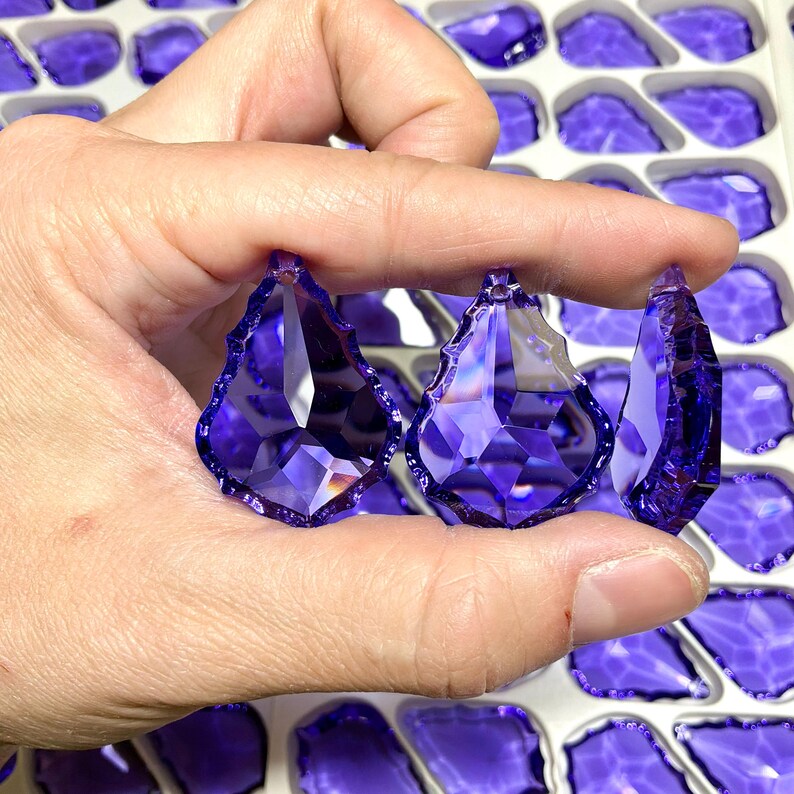 May include: Three faceted, teardrop-shaped, purple crystal pendants are held between fingers. The crystals have a pointed top and a jagged bottom edge. The background shows more purple crystal pendants in a white tray.