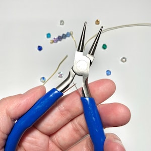 May include: Round nose pliers with blue handles are held in a hand. The pliers are silver and have long, tapered jaws. Small beads in various colors and a length of wire are scattered around the pliers, suggesting jewelry making.