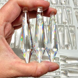 May include: Three clear crystal teardrop prisms with iridescent rainbow facets. The prisms are held in a hand and are approximately 3 inches long.