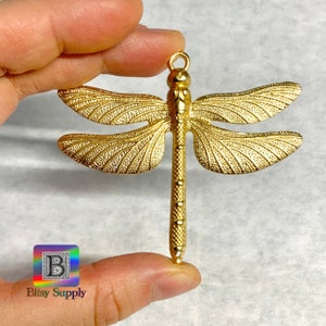 Puede incluir: Un colgante de libélula dorado con texturas detalladas en las alas y el cuerpo. La libélula está sujeta por una mano. El colgante tiene un lazo en la parte superior para colgar. El logotipo "Bitsy Supply" está en la esquina inferior izquierda.