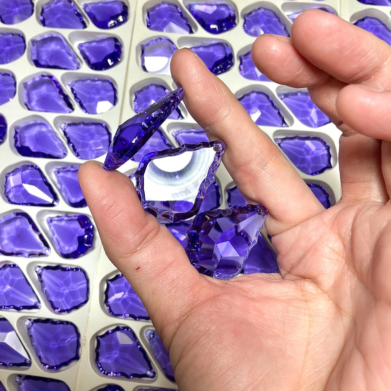 May include: Close-up of several faceted, deep purple glass pieces. Some pieces are held in a hand, while others are arranged in a white tray. The glass has a brilliant, reflective quality, suggesting they are decorative or for crafting.