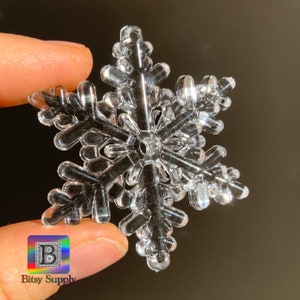 May include: A clear plastic snowflake ornament with a six-pointed star design. The snowflake is held in a hand.