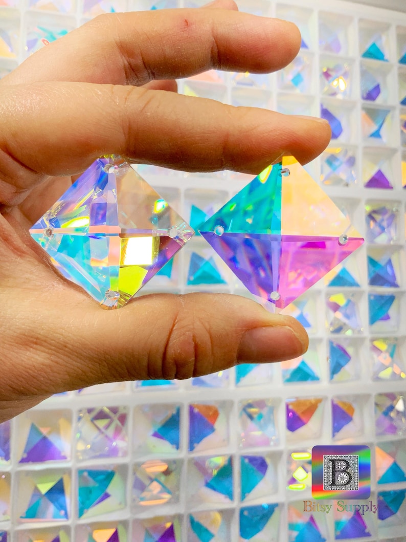 K&ouml;nnte beinhalten: Zwei regenbogenfarbene quadratische Glaskristalle mit einem Regenbogeneffekt. Die Kristalle werden in einer Hand vor einem Hintergrund aus vielen &auml;hnlichen Kristallen gehalten.