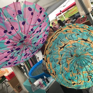 May include: Two colorful, hand-painted paper umbrellas with marbled designs. One umbrella is pink, blue, and purple with black dots. The other umbrella is teal, gold, and black.