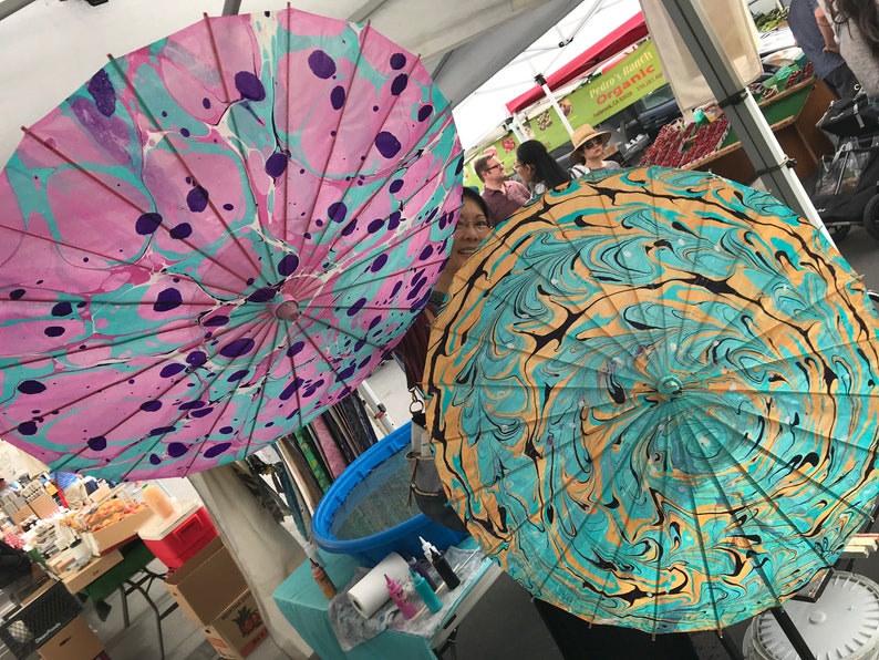 May include: Two colorful, hand-painted paper umbrellas with marbled designs. One umbrella is pink, blue, and purple with black dots. The other umbrella is teal, gold, and black.