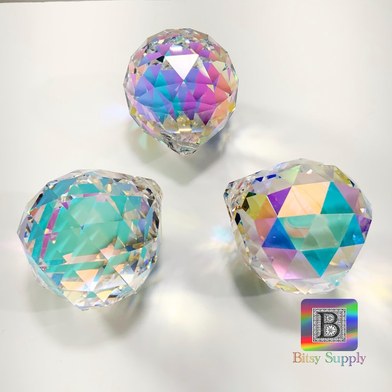 K&ouml;nnte beinhalten: Drei regenbogenfarbene Kristallkugeln mit facettierten Oberfl&auml;chen. Die Kugeln befinden sich auf einem wei&szlig;en Hintergrund.