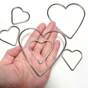 Puede incluir: Una colección de formas de corazón de alambre de color plateado en varios tamaños. Algunos corazones están dispuestos en un patrón anidado, mientras que otros están dispersos. Los corazones se muestran sobre un fondo blanco.