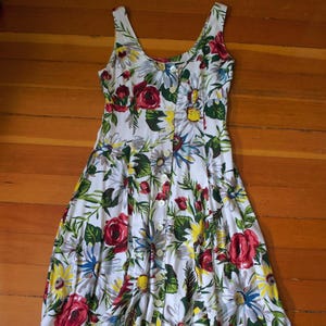 May include: White sleeveless dress with a floral pattern featuring red roses, yellow daisies, and blue accents. The dress has a button-down front and a flared skirt, perfect for a summer day. The dress is made of a lightweight fabric.