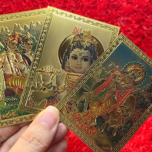 May include: Three rectangular gold-toned cards featuring embossed illustrations of Hindu deities. Each card displays a figure playing a flute, surrounded by intricate geometric borders. The metallic finish reflects light, highlighting the detailed relief work.