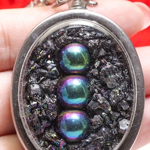 May include: Oval pendant with a silver-colored frame, featuring three iridescent spheres set against a backdrop of dark, crystalline fragments. The pendant is suspended from a silver chain.