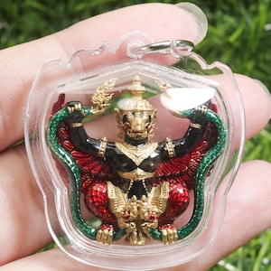 Könnte beinhalten: Ein Anhänger mit einer detaillierten, farbenfrohen Garuda-Figur. Die Figur hat einen schwarzen Körper, rote Flügel und goldene Akzente, umhüllt von einem transparenten Schutzgehäuse. Der Garuda hält eine grüne Schlange.