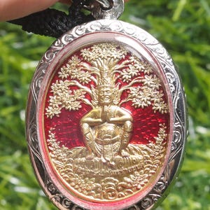 May include: An oval pendant with a silver-colored frame and a detailed gold-colored image of a seated figure with a tree-like design above. The background is a vibrant red. The pendant is suspended from a black cord.