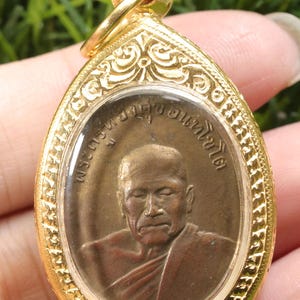 May include: Gold-colored pendant with an oval shape, featuring an image of a man in a brown robe. The pendant has intricate decorative patterns around the edge and a small loop at the top for a chain.
