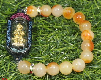 May include: A bracelet made of light orange and yellow beads with a gold charm in a black and blue frame. The bracelet is on a green grassy surface.