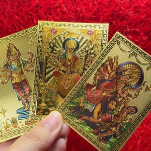 May include: Three rectangular gold-toned cards featuring embossed illustrations of Hindu deities. Each card displays intricate metallic detailing, including figures with multiple arms, ornate clothing, and lion motifs, all framed by a decorative border.
