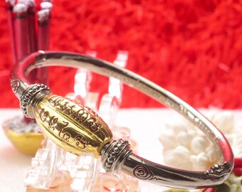 Puede incluir: Una pulsera rígida de tono plateado que presenta una cuenta central alargada de tono dorado con letras en relieve. La pulsera está acentuada con tapas decorativas plateadas con cuentas a ambos lados de la cuenta central y patrones de espirales grabados.
