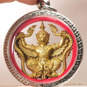 May include: A gold-coloured Garuda pendant encased in a circular silver-toned metal frame with intricate engravings. The pendant features a central figure of the mythical Garuda with wings spread, set against a vibrant red inner border lining.