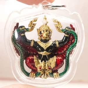 May include: A pendant featuring a detailed Garuda figure in black, gold, and red enamel, encased in a clear protective plastic shell. The mythical bird-like creature holds a green serpent in its hands, with intricate gold filigree details.