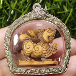 Op de afbeelding: Een gedetailleerde hanger met een gebeeldhouwd, crèmekleurig mythisch wezen in een heldere, gewelfde behuizing. De hanger is ingekapseld in een zilverkleurig, sierlijk frame met een lus om op te hangen. De achtergrond is een vage groene kleur.