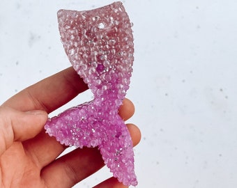 Pode incluir: Um amuleto de cauda de sereia rosa e prateado feito de resina com acabamento brilhante.