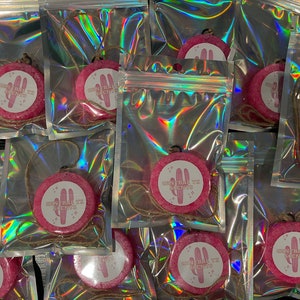 May include: Multiple pink round car air fresheners with a white cactus design and the text "Desert Sun Scents" hanging from a string in clear plastic bags.
