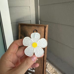 May include: White daisy-shaped ornament with a yellow center and a black loop for hanging. The ornament has a textured surface and is held in front of a wooden sign.