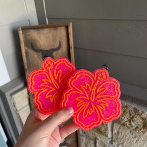 May include: Two pink hibiscus flower shaped coasters with orange glitter outlines. The coasters are held in a hand.