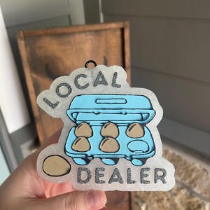 May include: A light blue egg carton-shaped ornament with the words "LOCAL DEALER" in silver glitter. The ornament features six brown eggs inside the carton and one egg outside.