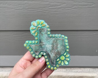May include: A turquoise-colored Texas-shaped decorative item with a textured surface and a border of green and yellow floral accents. The item has a rustic, weathered appearance, and is likely a handcrafted piece.