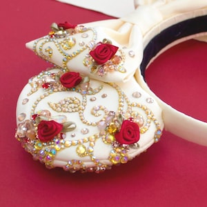 May include: Ivory-coloured headband with a decorative bow and embellishments. The bow features red roses, gold-coloured swirls, and sparkling crystals. The headband has a black band on the inside.
