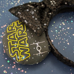 Może przedstawiać: Czarna opaska do włosów z cekinami, z czarną kokardką z tkaniny i czarnym uchem z tkaniny z napisem "STAR WARS" w żółtych kryształach i srebrnym kryształowym X-Wingiem.