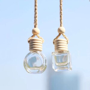 May include: Two glass car air freshener bottles with wooden stoppers and beige string hangers. The bottle on the left is round and the bottle on the right is square.