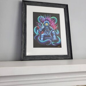 May include: Framed art print featuring a diver seated on a throne of tentacles, holding a spear. The diver is in a blue diving suit with a yellow helmet. The artwork has a black background with vibrant teal, purple, and pink accents. The frame is black.