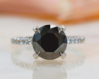 May include: A silver ring featuring a large, round, black gemstone as the focal point. The band is adorned with smaller, clear gemstones, adding sparkle. The ring is set against a soft, blurred background, highlighting the central black stone.