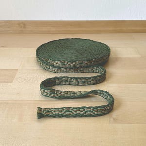 Può includere: Un rotolo avvolto di nastro in tessuto intrecciato in una tonalità verde bosco profondo. Il nastro presenta un motivo geometrico ripetitivo nei toni del verde chiaro, arancione e verde scuro, con un aspetto intrecciato e testurizzato.