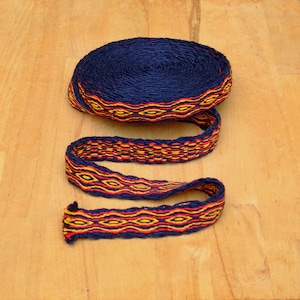 Peut inclure: Un rouleau de galon tissé bleu foncé avec un motif répétitif jaune, orange et rouge. Une longueur du même galon est déroulée, montrant le motif détaillé. Le galon est probablement utilisé pour l'artisanat ou la décoration.
