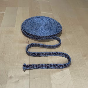 May include: A coiled roll of blue and burgundy woven trim sits next to a length of the same trim, uncoiled. The trim has a repeating geometric pattern in burgundy against a blue background. The trim is on a light brown wooden surface.