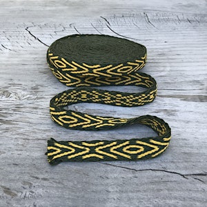 May include: A green and yellow woven ribbon with a geometric pattern. The ribbon is rolled up and lying on a wooden surface.