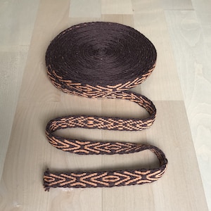 May include: A brown woven ribbon with an orange geometric pattern. The ribbon is rolled up and laid out flat on a wooden surface.