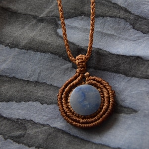 Pode incluir: Um colar de macramé castanho com um pendente redondo de pedra azul clara. O pendente é circundado por uma espiral de cordão castanho atado. O cordão do colar também é castanho e trançado.