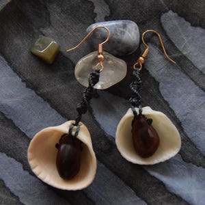 Könnte beinhalten: Handgefertigte Ohrringe mit natürlichen Muscheln, dunkelbraunen Samenkapseln und goldfarbenen Ohrhaken. Die Ohrringe hängen an schwarzen, gedrehten Kordeln. Im Hintergrund befinden sich kleine Steine auf einer strukturierten blau-grauen Oberfläche.
