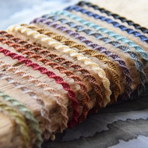 Könnte beinhalten: Eine Sammlung von 20 bunten geflochtenen Schnüren, die in einer Reihe auf einer Holzoberfläche angeordnet sind. Die Schnüre sind aus einem weichen, natürlichen Material gefertigt und in verschiedenen Farben erhältlich, darunter Schwarz, Blau, Grün, Braun, Rot und Weiß.