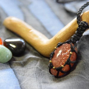 Può includere: Primo piano di un ciondolo in pietra di sole avvolto in un cordoncino nero con perline dorate. Il ciondolo è appoggiato su una superficie di legno e uno sfondo di tessuto grigio. Ci sono altre tre pietre nell'immagine: una pietra rossa, una pietra verde e una pietra nera.