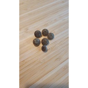 May include: A collection of six circular metal buttons featuring a detailed, raised sunburst or floral pattern with small, beaded textures radiating from the centre. The buttons have a dark, oxidised silver finish with highlights on the raised edges.