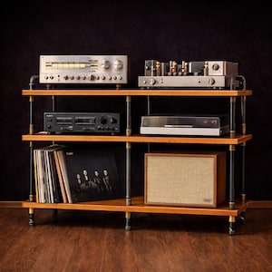 May include: A wooden audio rack with three shelves, holding vintage stereo equipment. The rack has black metal pipe supports and castors. Components include a receiver, amplifier, cassette deck, turntable, speaker, and vinyl records. The speaker has a tan fabric front.