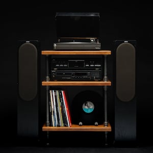 May include: A black turntable sits on a wooden shelf unit with two black speakers on either side. The shelf unit has three shelves, the top shelf holds the turntable, the middle shelf holds a CD player and a cassette player, and the bottom shelf holds a stack of vinyl records and a black vinyl record.