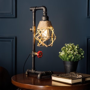 May include: Industrial-style table lamp with a black pipe frame and a geometric shade. The shade is wrapped with natural rope and features a red valve detail. A small potted plant and books are on the table.