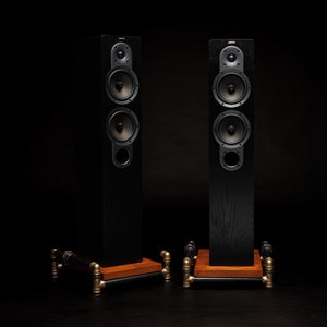 May include: Two tall, black speakers with multiple circular drivers and the brand name "Jamo" at the top. The speakers sit on wooden platforms supported by a network of black and gold pipes. The background is a solid black.