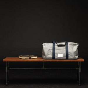 May include: A grey canvas duffel bag with black straps sits on a wooden bench with a brown leather-bound book. The bench has black metal legs.